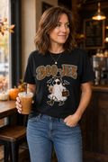 A woman wearing a black graphic t-shirt with a Halloween ghost design and the text 'BOO-JEE' in gold and orange, paired with blue jeans, standing in a cozy cafe environment and holding an iced coffee.