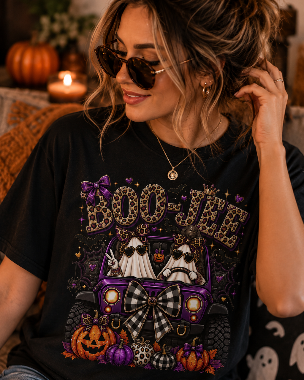 A woman wearing a black graphic t-shirt with a Halloween-themed design featuring a purple jeep, two ghosts, pumpkins, and the text 'BOO-JEE' in leopard print. She is wearing sunglasses and a necklace in a cozy, warmly lit indoor setting.
