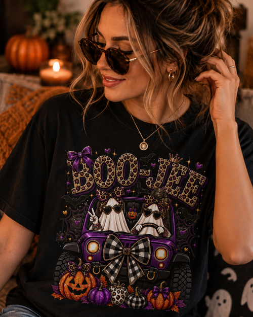 A woman wearing a black graphic t-shirt with a Halloween-themed design featuring a purple jeep, two ghosts, pumpkins, and the text 'BOO-JEE' in leopard print. She is wearing sunglasses and a necklace in a cozy, warmly lit indoor setting.