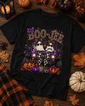 A black short-sleeve graphic T-shirt laid flat on a rustic wooden surface with autumn props. The shirt features a Halloween-themed print with the words “BOO JEE” in leopard-print letters above a purple jeep carrying two ghosts, surrounded by pumpkins, spiderwebs, and fall leaves.