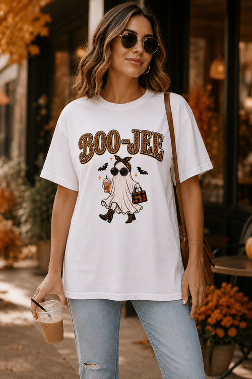 A woman wearing a white graphic t-shirt with a 'BOO-JEE' ghost and cat Halloween design, paired with light blue jeans and sunglasses, holding an iced coffee in an outdoor cafe setting.