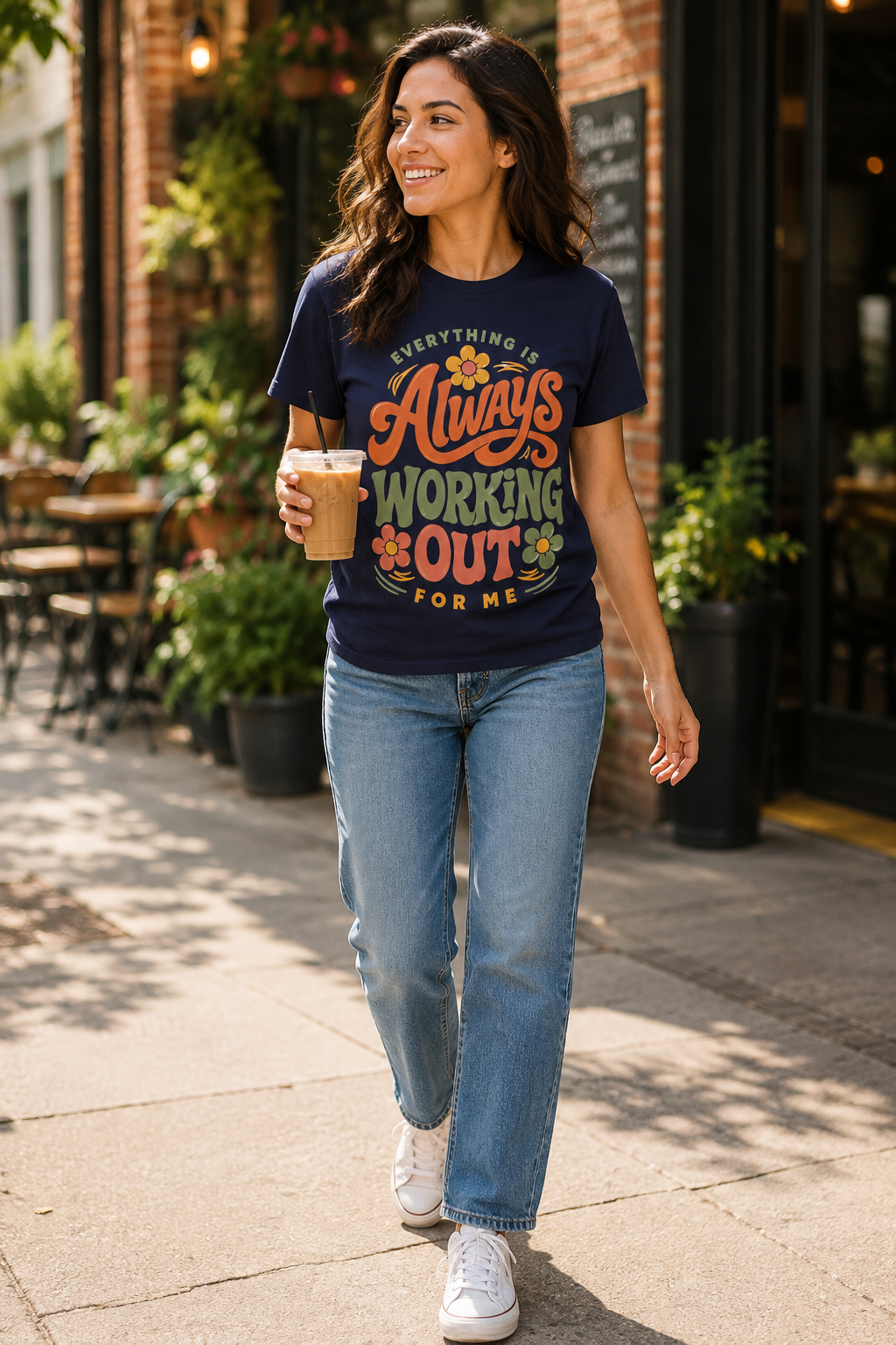 A woman walking on a sunny sidewalk wearing a navy blue graphic t-shirt with the text 'EVERYTHING IS Always WORKING OUT FOR ME' in orange and yellow lettering, paired with light-wash blue jeans and white sneakers.