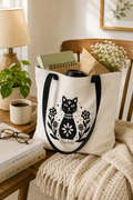 A cream canvas tote bag with black handles and a black cat-and-floral graphic sits on a wooden chair in a cozy room setting, filled with notebooks, flowers, and a bottle.