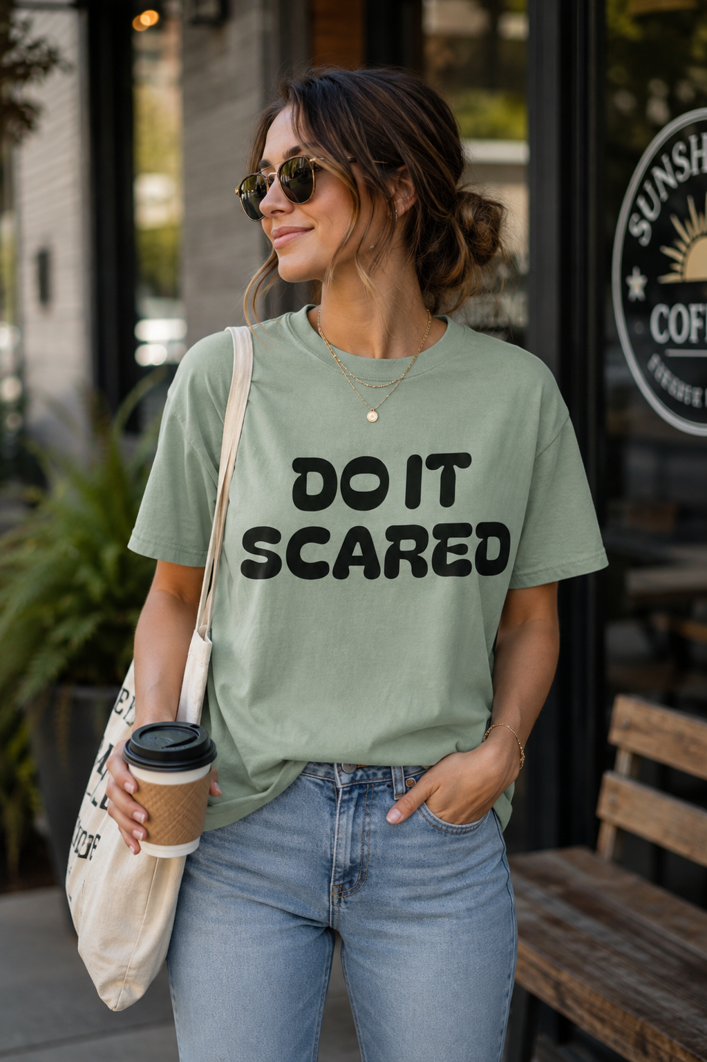 A woman wearing a sage green graphic t-shirt with the text 'DO IT SCARED' in black, paired with light blue jeans and carrying a white tote bag, standing outside a coffee shop.