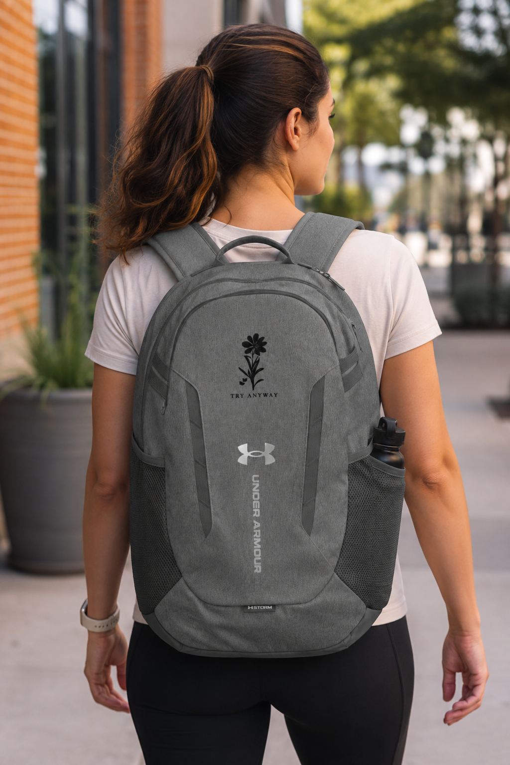 A woman viewed from behind wearing a gray Under Armour backpack on a paved outdoor walkway.