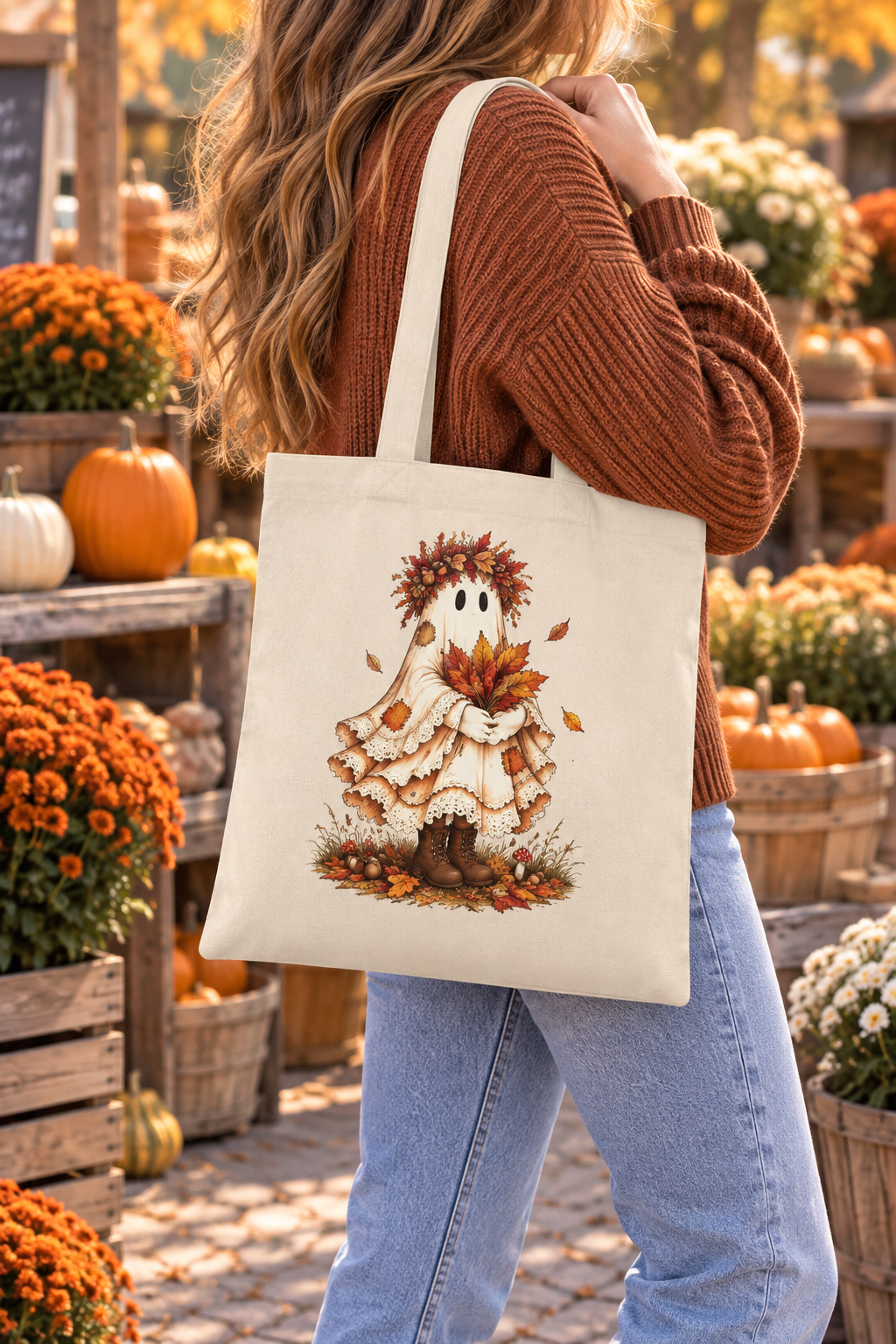 A woman wearing a rust-brown ribbed sweater and light blue jeans carries a cream canvas tote bag with a whimsical autumn ghost illustration. The background is a rustic fall market scene filled with orange pumpkins, mums, and wooden crates.