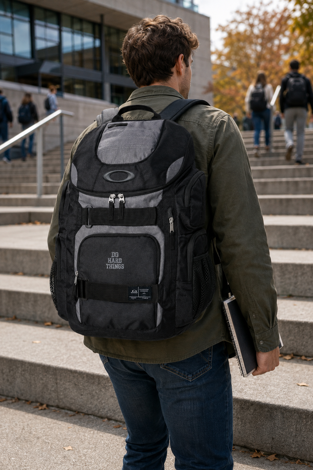 A man viewed from behind wearing a black and gray Oakley backpack while standing on outdoor concrete steps near a modern building.