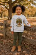 A young girl with dark curly pigtails stands outdoors in a wooded park setting wearing a light gray crewneck sweatshirt with a 'Every Night Owl Becomes an Auburn' tree graphic, olive green leggings, and tan ankle boots.