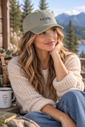 A woman sitting outdoors by a lake with mountains and pine trees in the background, wearing an olive green baseball cap with blue embroidered text that reads 'keep going.' She is also wearing a cream knit sweater and blue jeans.