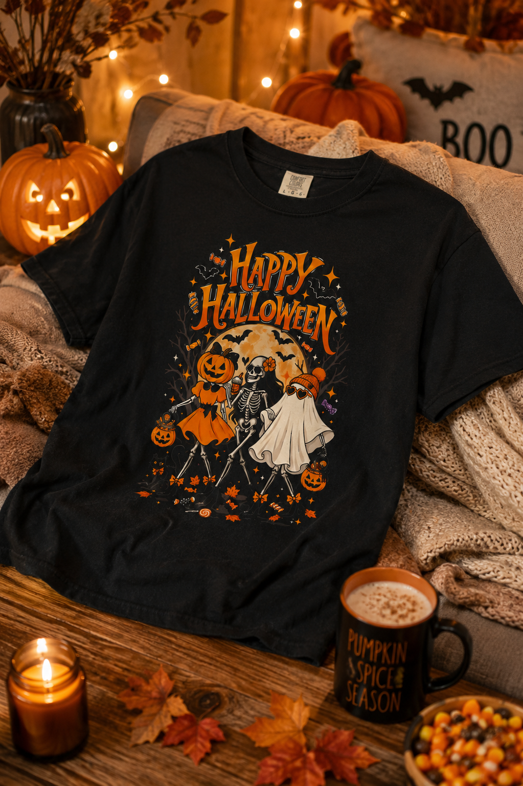 A black graphic t-shirt featuring a 'Happy Halloween' skeleton and ghost illustration, styled on a cozy couch with pumpkins, a candle, and a mug in a warm autumn setting.