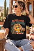 Woman wearing a black “I’m Not About This Weather” graphic T-shirt featuring a funny overheated sun and El Niño design while relaxing on a sunny desert patio with cacti.