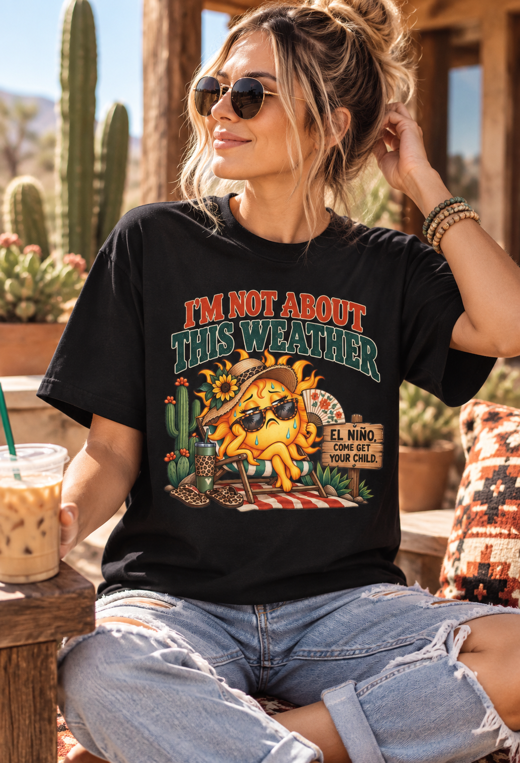Woman wearing a black “I’m Not About This Weather” graphic T-shirt featuring a funny overheated sun and El Niño design while relaxing on a sunny desert patio with cacti.