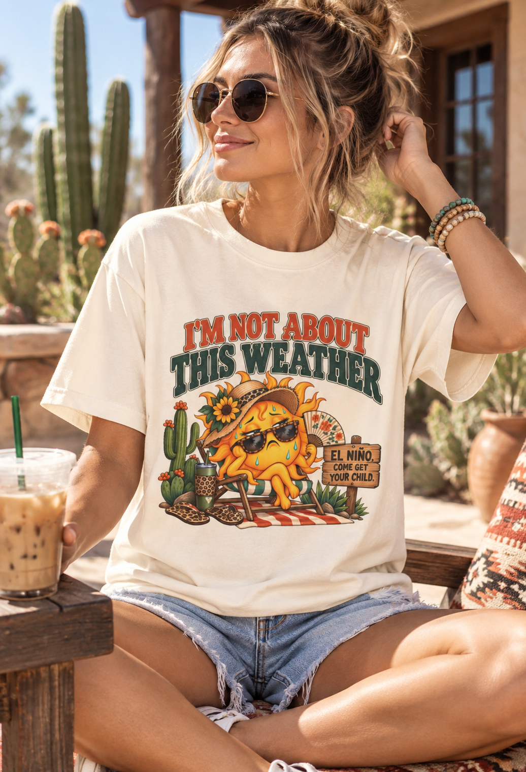Woman wearing a cream “I’m Not About This Weather” funny summer graphic T-shirt with retro sun and El Niño design in a desert cactus setting.