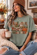 A woman wearing a sage green graphic t-shirt with a 'SWEATER WEATHER' autumn design, sitting in a cozy room and holding a mug.