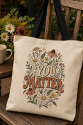 A beige canvas tote bag with black handles, featuring a 'YOU MATTER' floral graphic, resting on a rustic wooden chair next to a mug of coffee with flowers in the background.