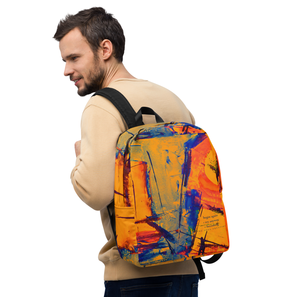A man wearing a beige sweatshirt and dark jeans is shown from the back and side, carrying a backpack with a vibrant abstract paint-splatter design in orange, yellow, blue, and red on a plain white background.