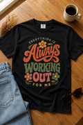 Black graphic T-shirt featuring the phrase “Everything Is Always Working Out for Me” in retro terracotta, sage green, dusty rose, and mustard lettering, styled with light-wash jeans, sunglasses, dried flowers, and coffee on a warm wood table.