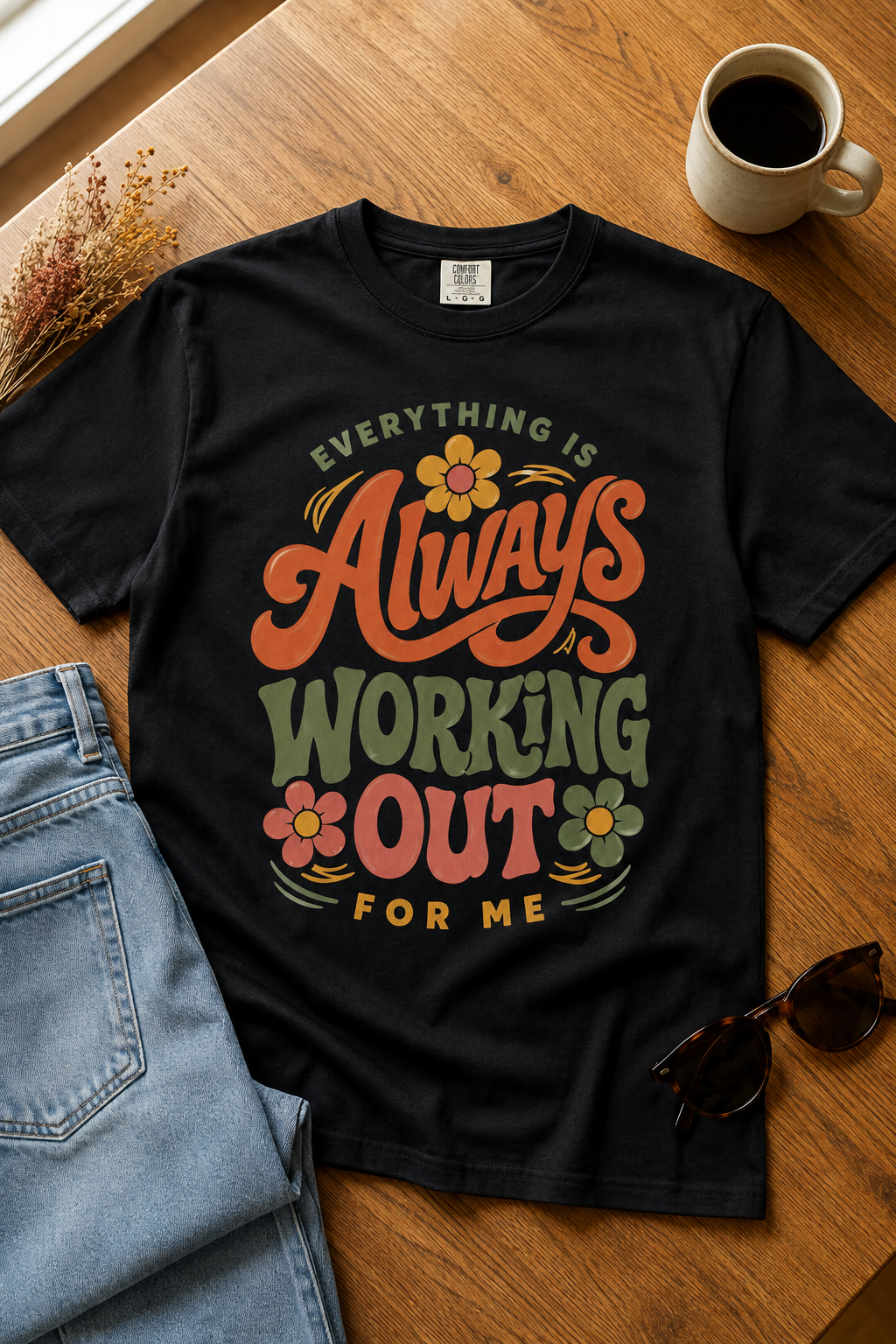 Black graphic T-shirt featuring the phrase “Everything Is Always Working Out for Me” in retro terracotta, sage green, dusty rose, and mustard lettering, styled with light-wash jeans, sunglasses, dried flowers, and coffee on a warm wood table.