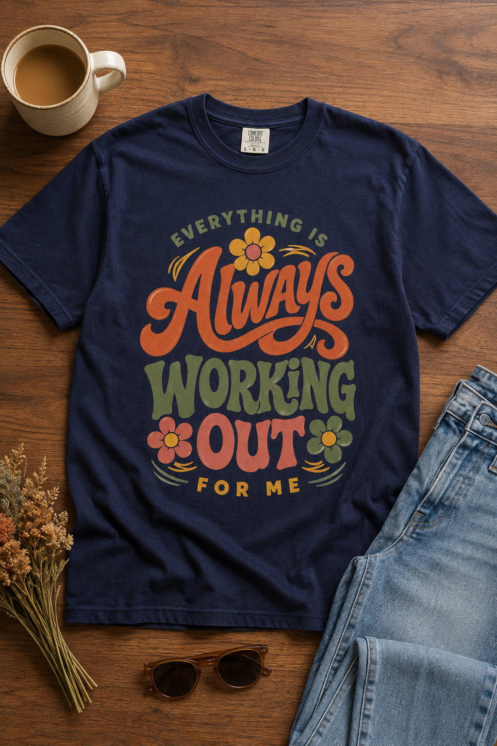 A navy blue graphic t-shirt laid flat on a wooden surface, styled with a coffee mug, sunglasses, dried flowers, and folded blue jeans. The t-shirt features a retro-style colorful graphic that reads 'EVERYTHING IS Always WORKING OUT FOR ME' with floral accents.