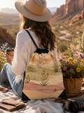 A woman in a wide-brim straw hat sits on a blanket in a desert canyon setting, holding a coffee cup and wearing a drawstring backpack with a printed landscape design. A basket of flowers and a camera are nearby.